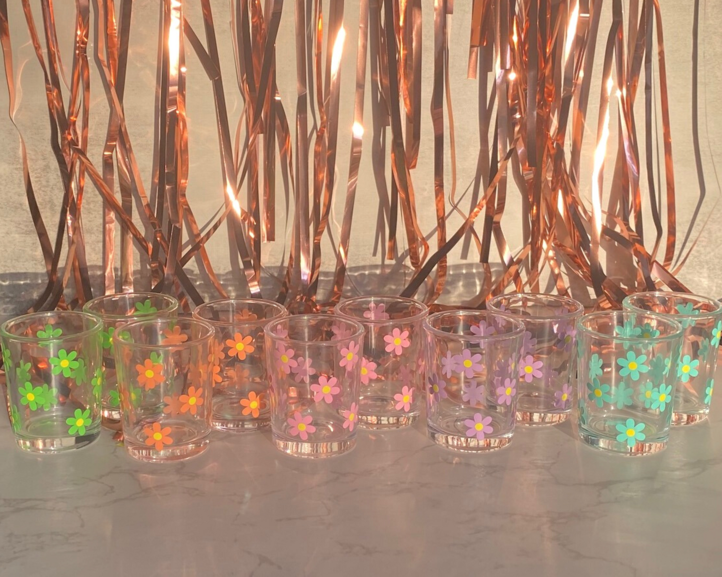 Daisy Flower Shot Glass, shown in the colors of the rainbow, pink, orange, green, mint, blue, and white, with a fun party background