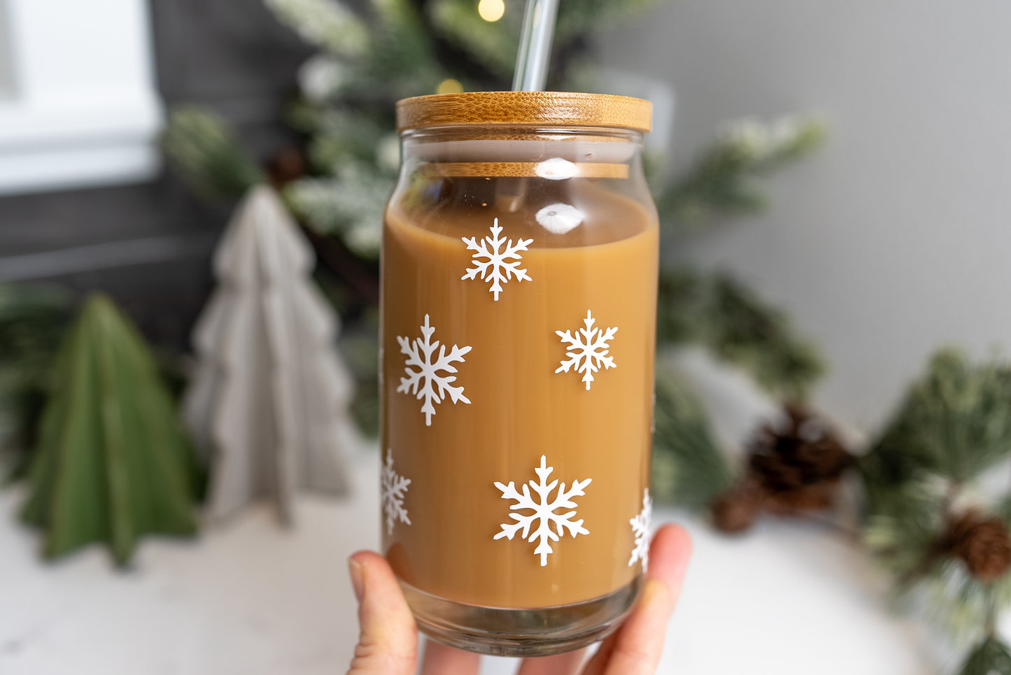 iced coffee glass cup with white snowflakes on it, pictured with a bamboo lid and clear glass straw with a christmas decor background