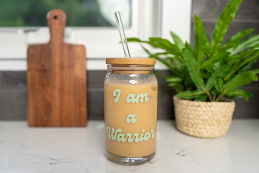 I am a Warrior Glass Cup in retro font in pastel green with bamboo lid and clear glass straw, filled with iced coffee on a minimalist background with green plant, 16oz beer can glass