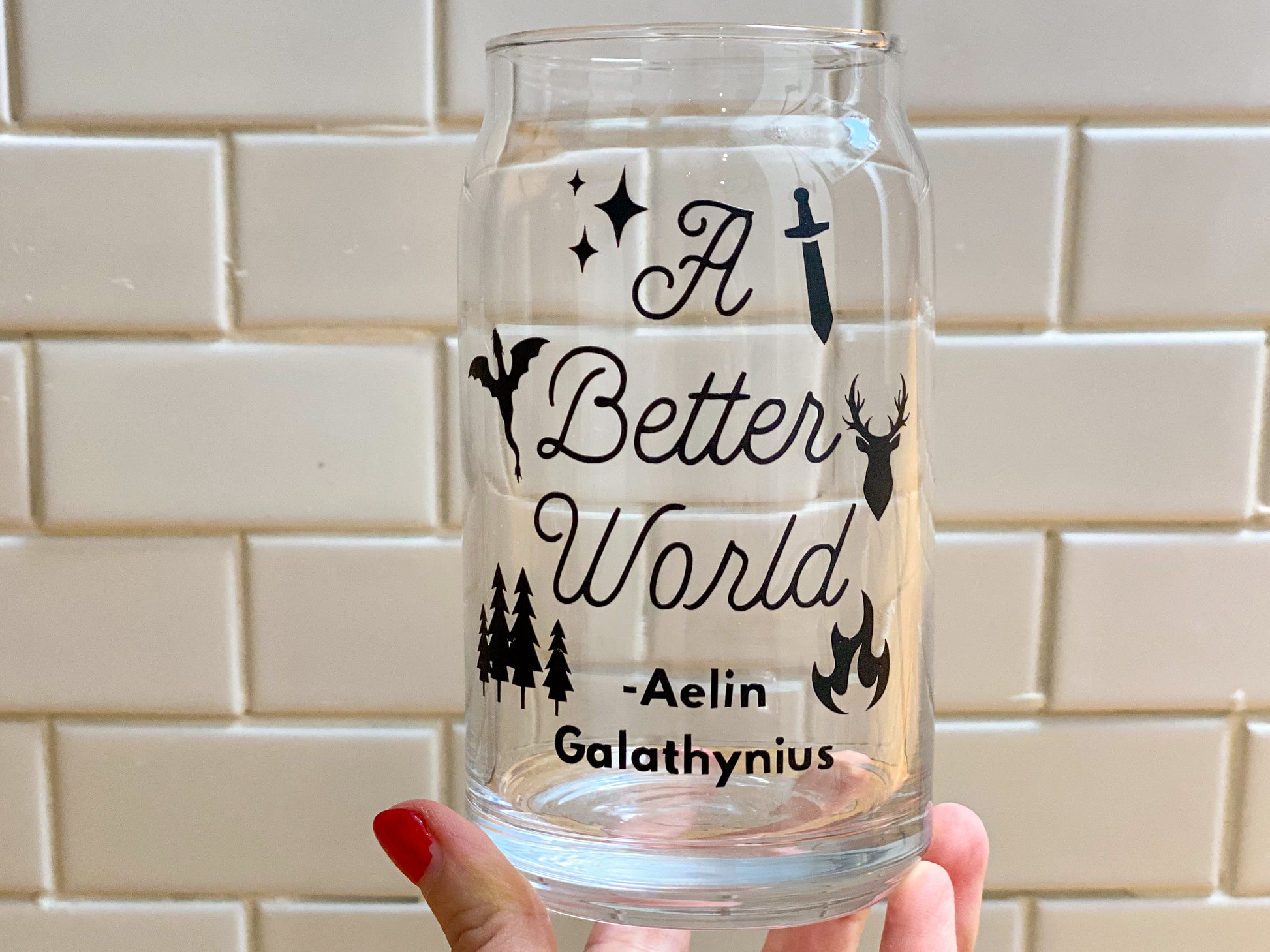 Glass cup for throne of glass book lovers with the quote from aelin galathynius that says a bettwe rold with 6 icons of stars, sword, wyrven, stag, pine trees, and fire on it, all in a black font, white subway tile background with cup in hand