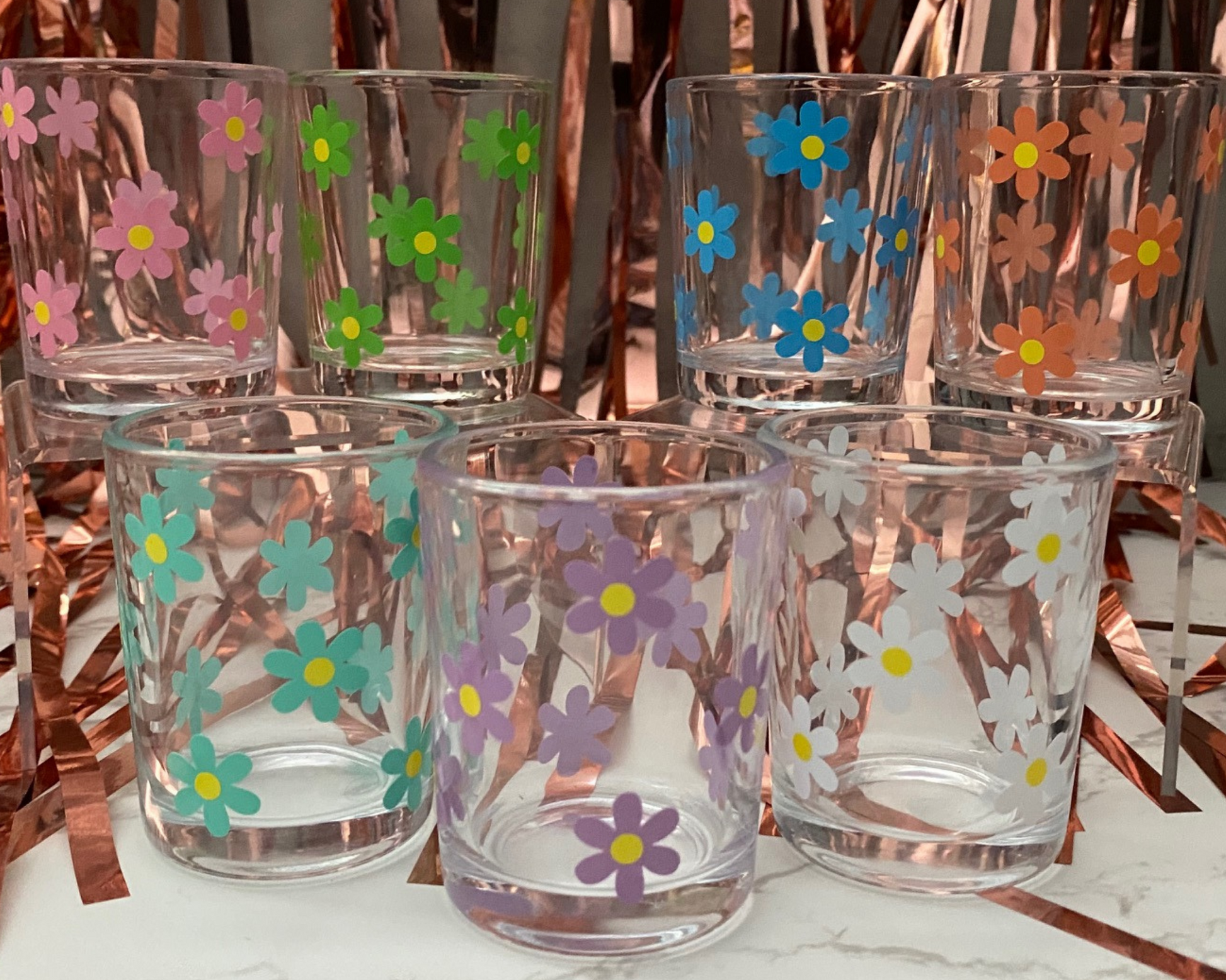seven shot glasses with a daisy flower design on it in green, mint, blue, orange, purple, pink and white with yellow dots in the middle, fun party background