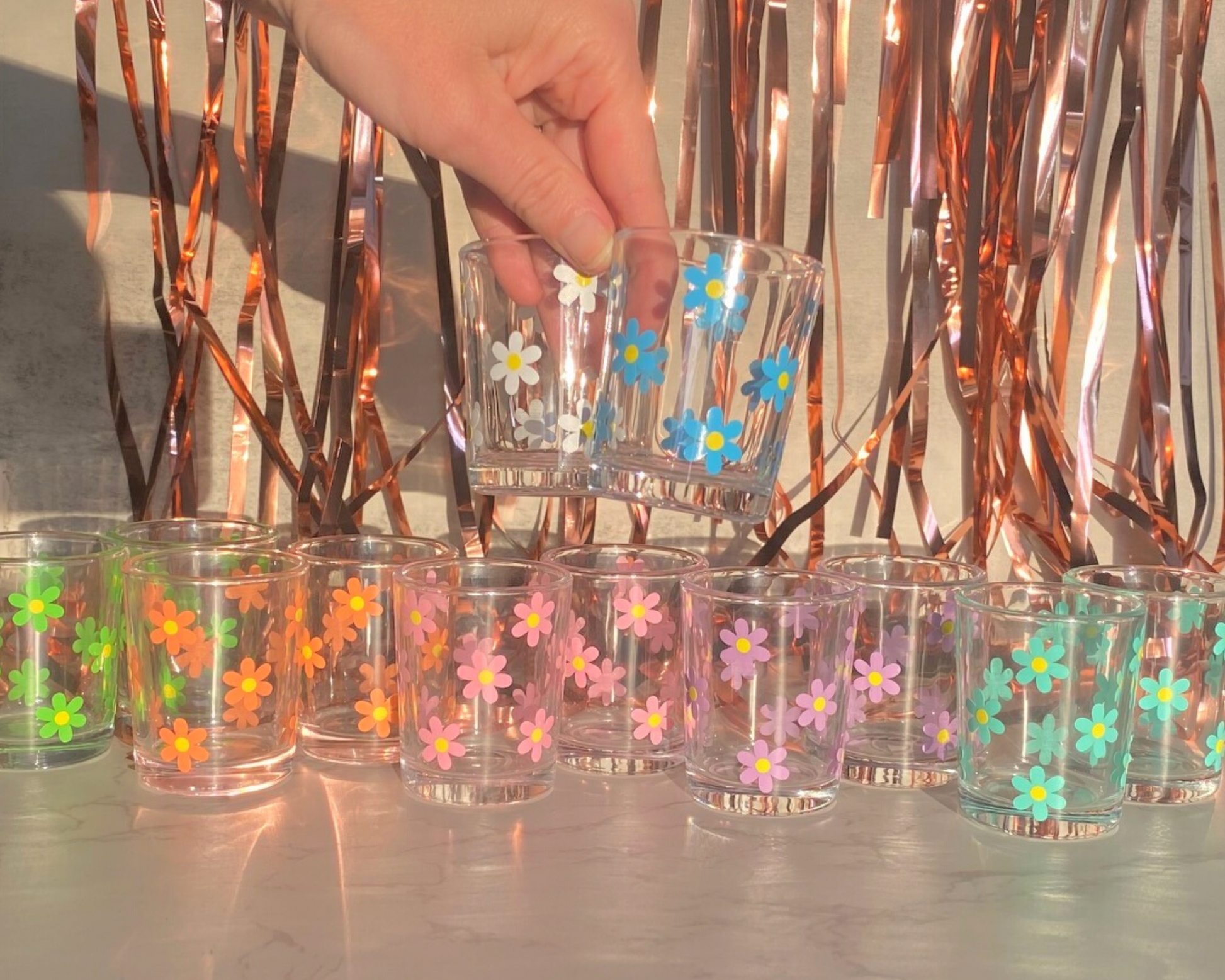 Daisy Flower Shot Glass, shown in the colors of the rainbow, pink, orange, green, mint, blue, and white, with a fun party background