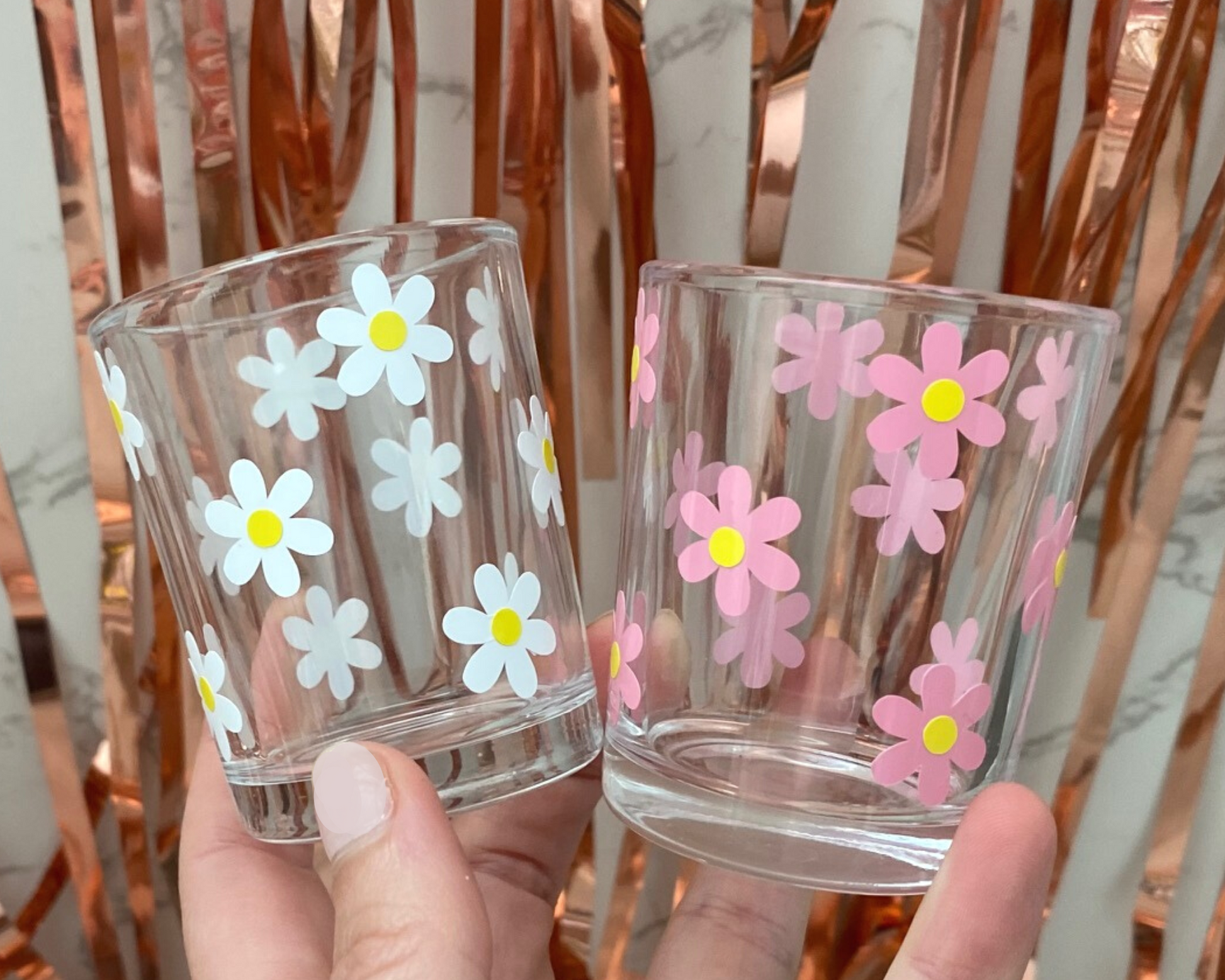 2 shot glasses in someones hand with a daisy flower design on it in pink and white with yellow dots in the middle, fun party background