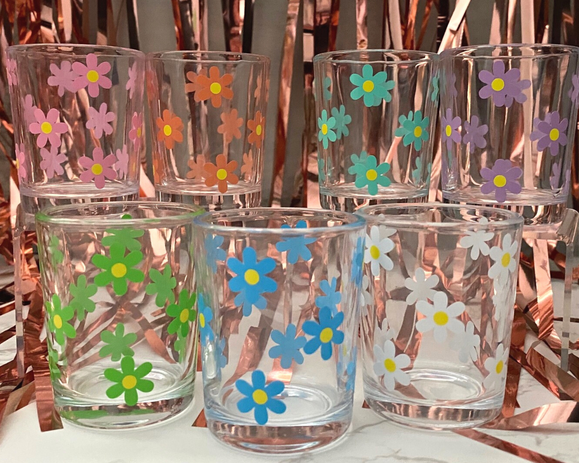 seven shot glasses with a daisy flower design on it in green, mint, blue, orange, purple, pink and white with yellow dots in the middle, fun party background