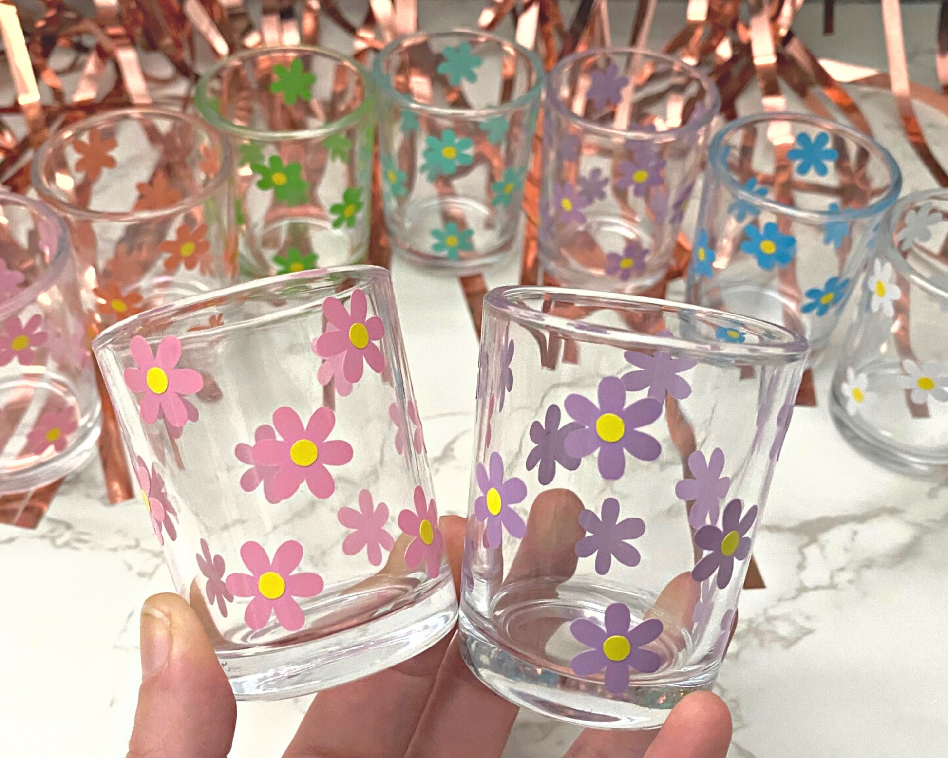 2 shot glasses in someones hand with a daisy flower design on it in pink and white with yellow dots in the middle, fun party background